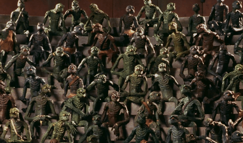 A crowd of reptile-aliens sit above the arena, watching the combat below